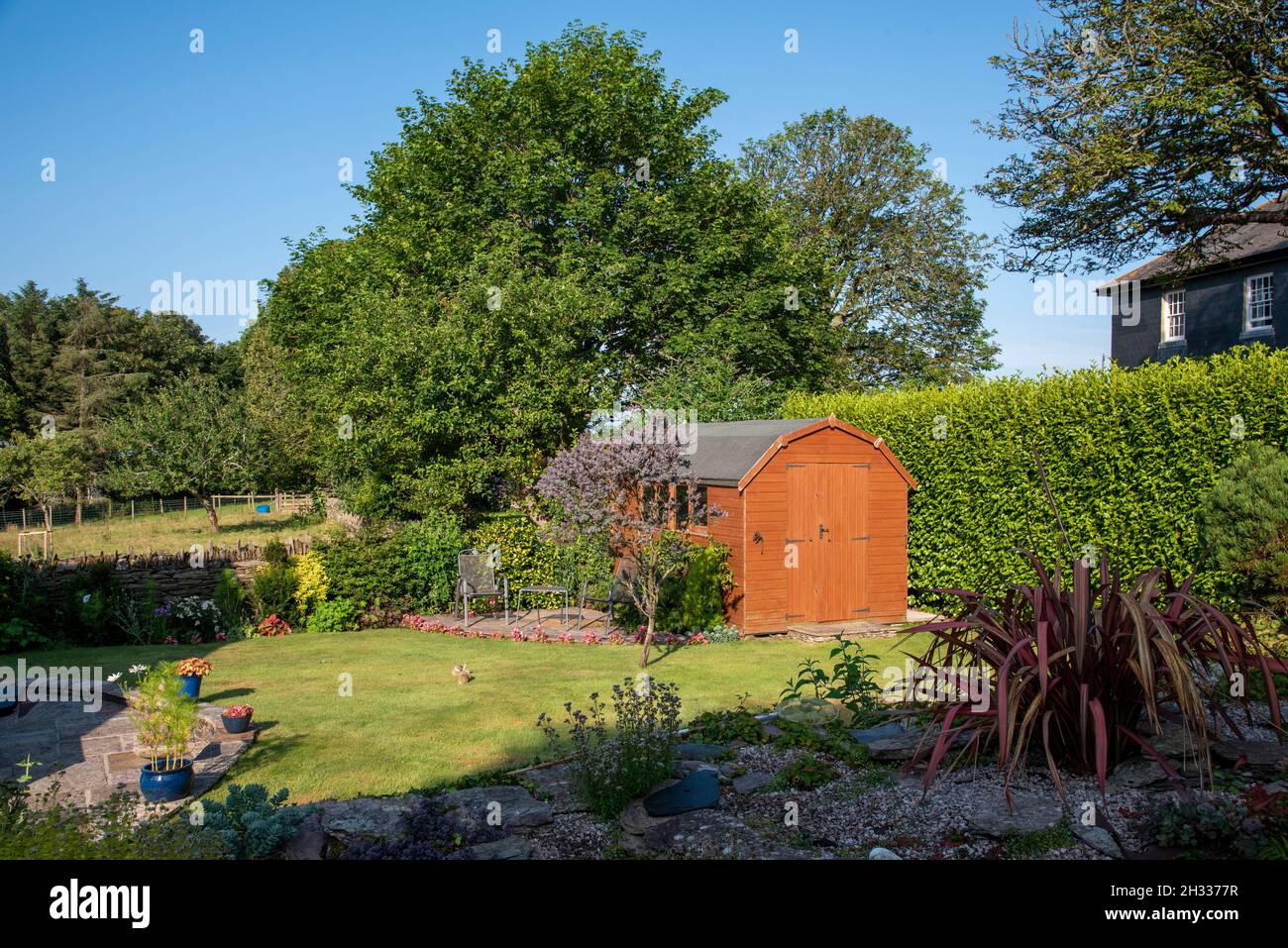 Devon, England, UK. 2021.  A Dutch barn style garden shed with double doors standing with patio, tables and chairs in an English country garden. Stock Photo