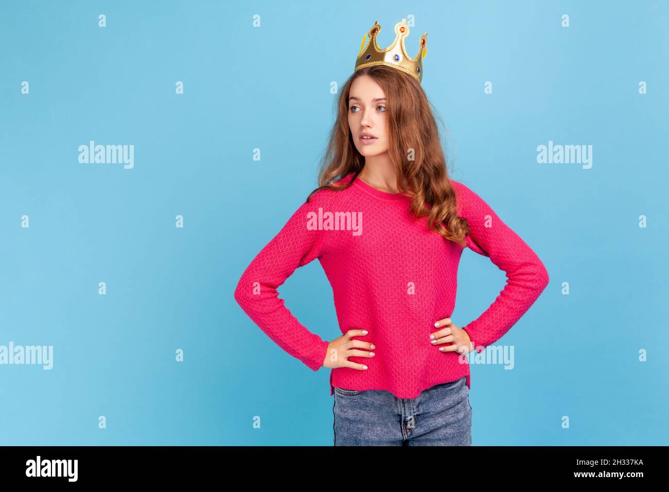 Arrogant woman wearing pink pullover, in crown on head looking away with confident expression, self-motivation and dreams to be best. Indoor studio shot isolated on blue background. Stock Photo