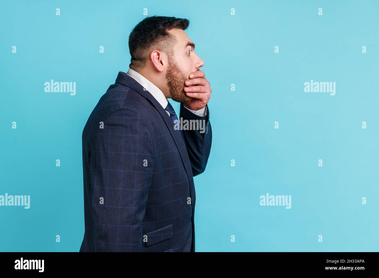 I won't say anyone. Side view of bearded man wearing official style suit covering mouth with hand, keeping terrible secret truth, don't want to talk. Indoor studio shot isolated on blue background. Stock Photo