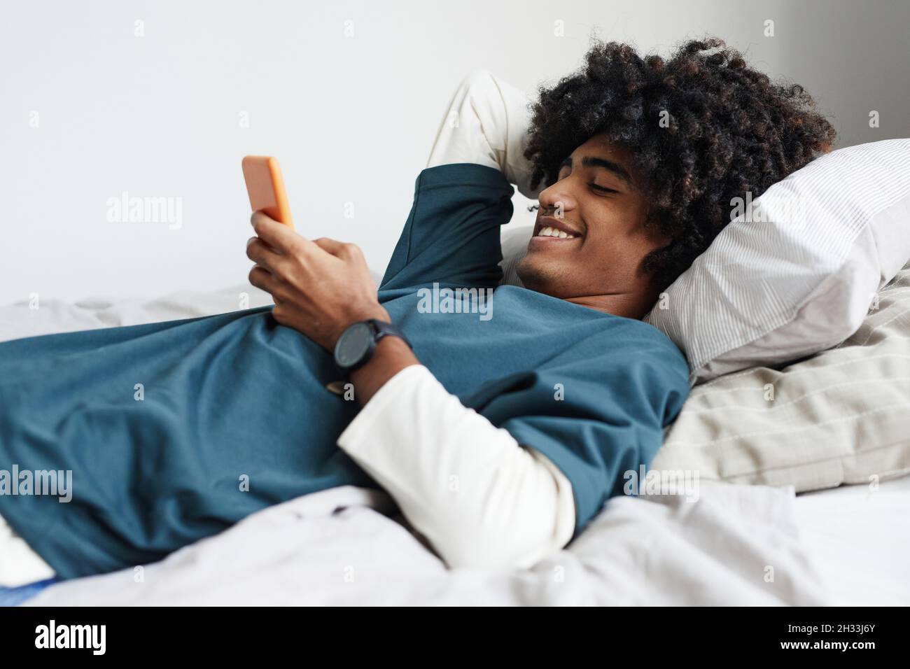 Side view portrait of young African-American man lying on bed at home and smiling while using smartphone Stock Photo