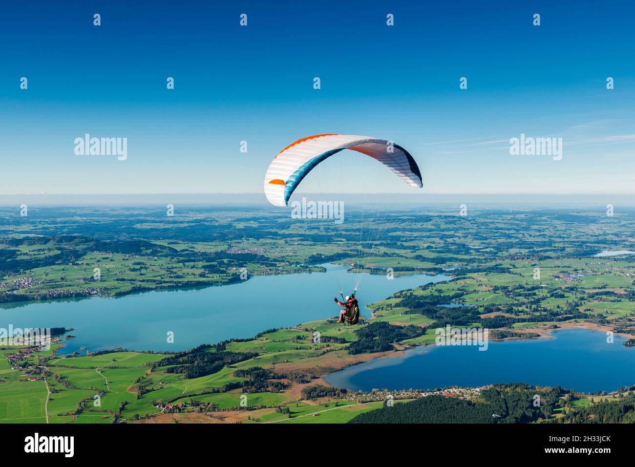 Paraglider pilots over a lake landscape in Bavaria in front of a blue sky and a wonderful panoramic view. Stock Photo
