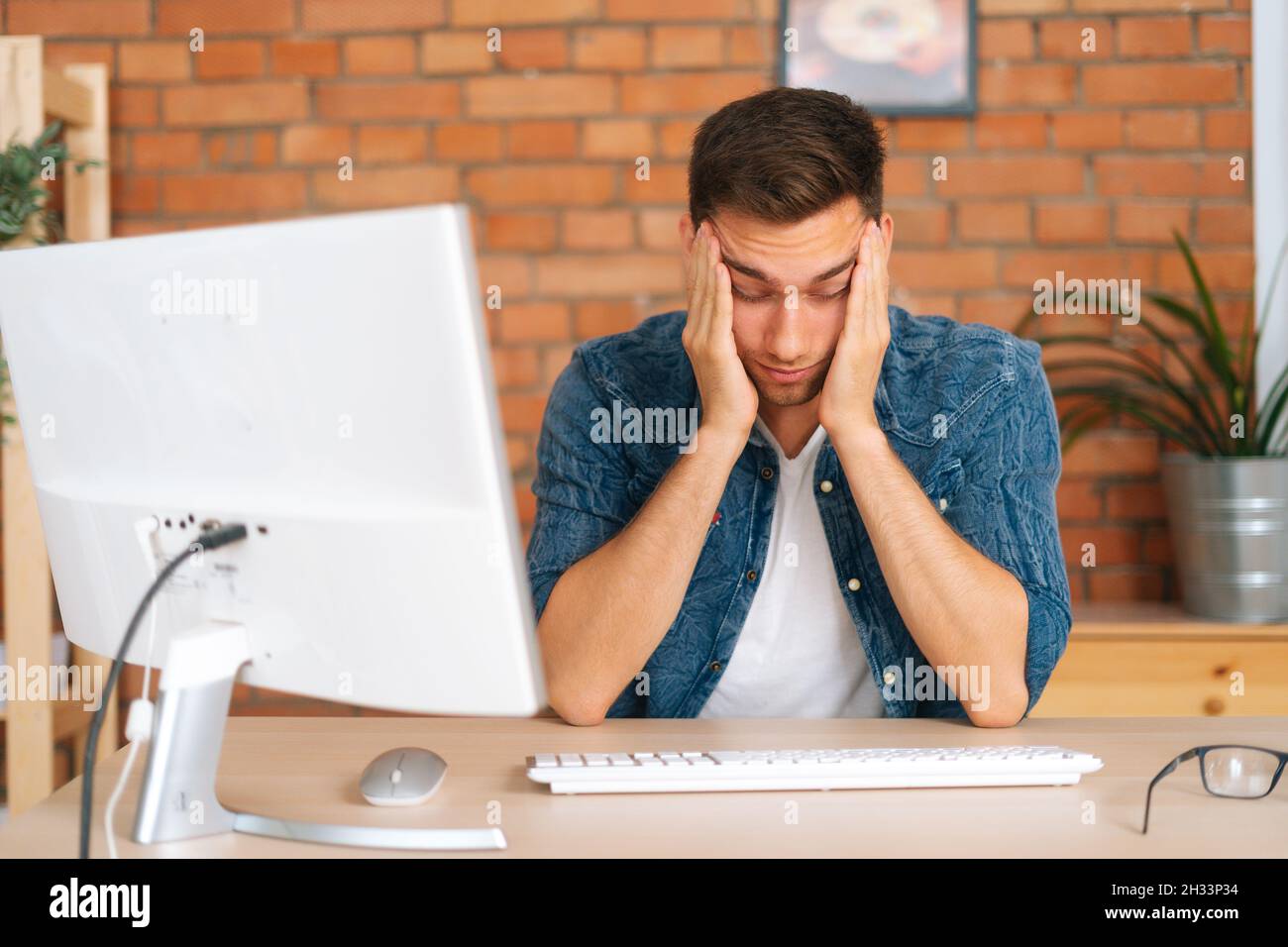 Portrait of frustrated exhausted young man touching temples feeling unbearable headache sitting at desk with desktop computer at home office. Stock Photo