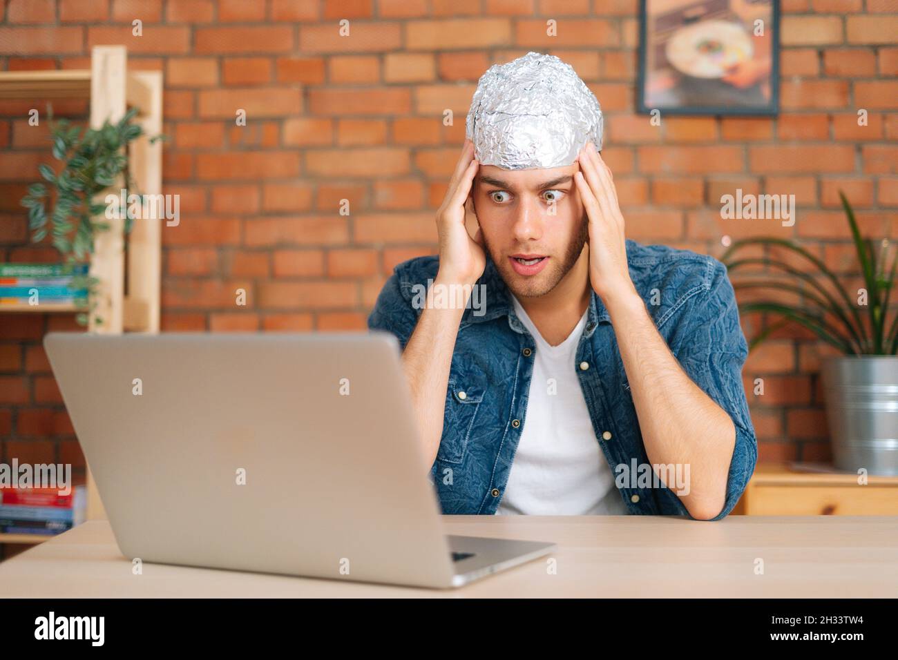 Portrait of scared afraid male conspiracy theorist wearing aluminum foil protect brain watching shocked online video content using laptop computer. Stock Photo