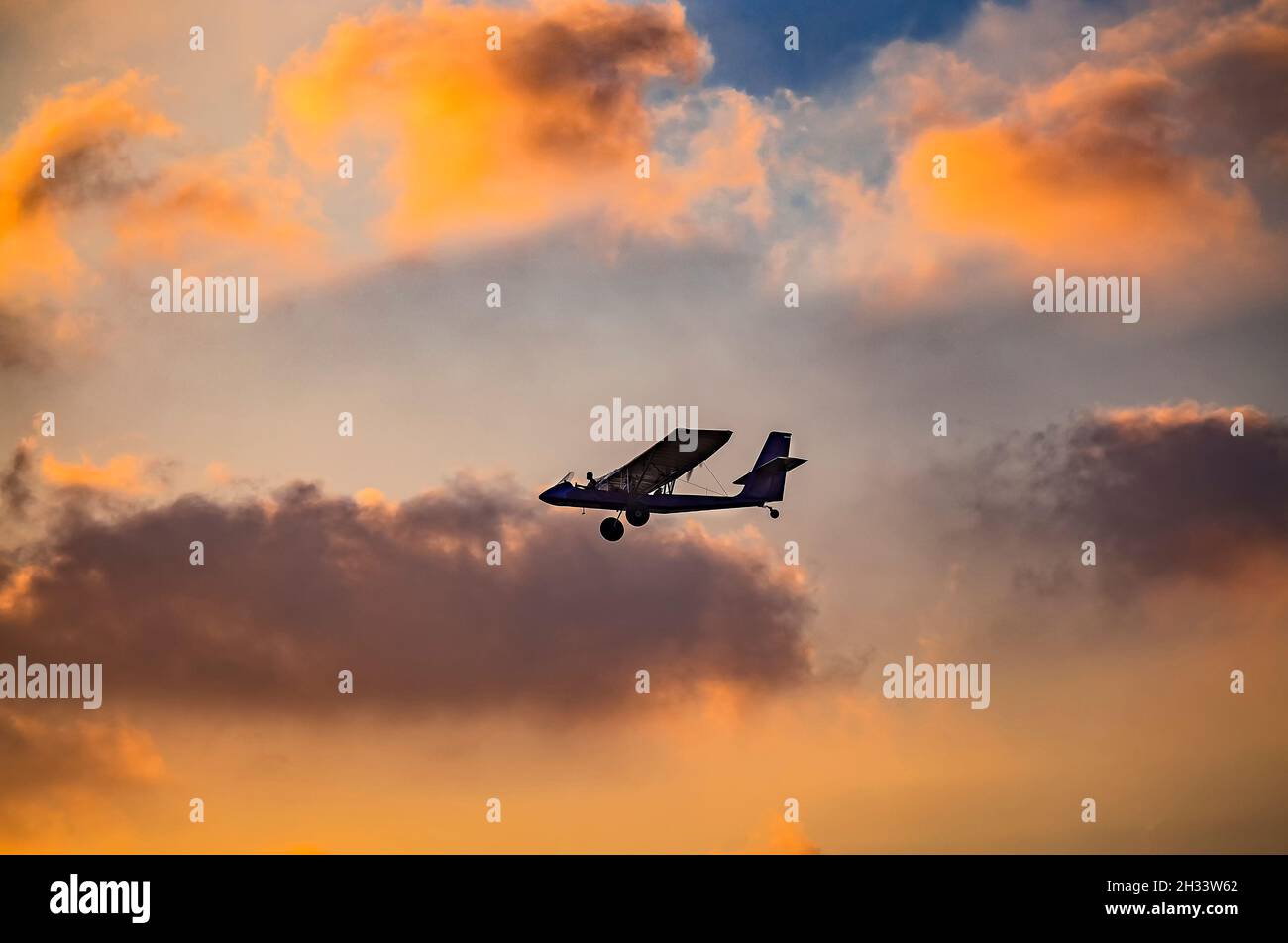 silhouette of ultralight microlite aircraft flying with a pilot and a passenger against sunset sky. Stock Photo