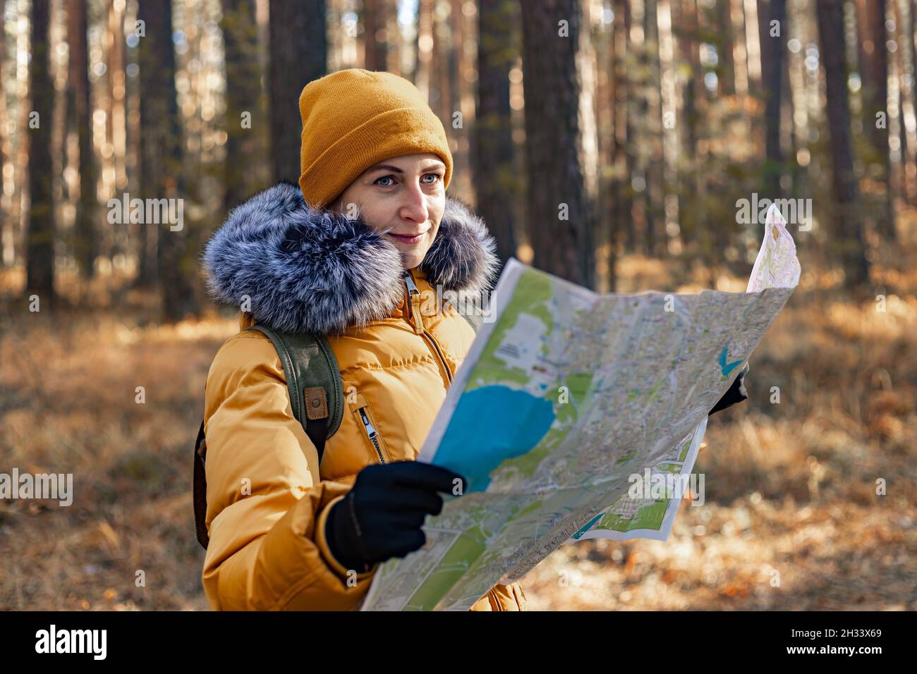 Young smiling woman in a warm orange jacket. Attractive Caucasian ethnicity woman walks in the park with a backpack. She looks at the map. Hiking in t Stock Photo