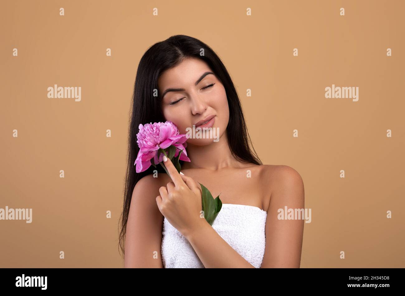 Tender armenian woman holding beautiful peony flower and closing eyes, posing over beige studio background. Portrait of lady having silky skin and nud Stock Photo