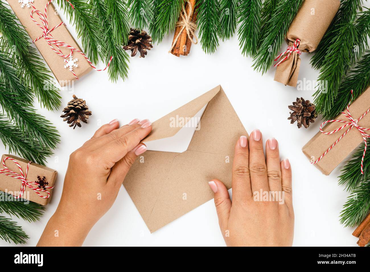 top view of woman hands sealing craft envelope with letter for Santa Claus in frame of Christmas tree branches, gift boxes on white background Stock Photo