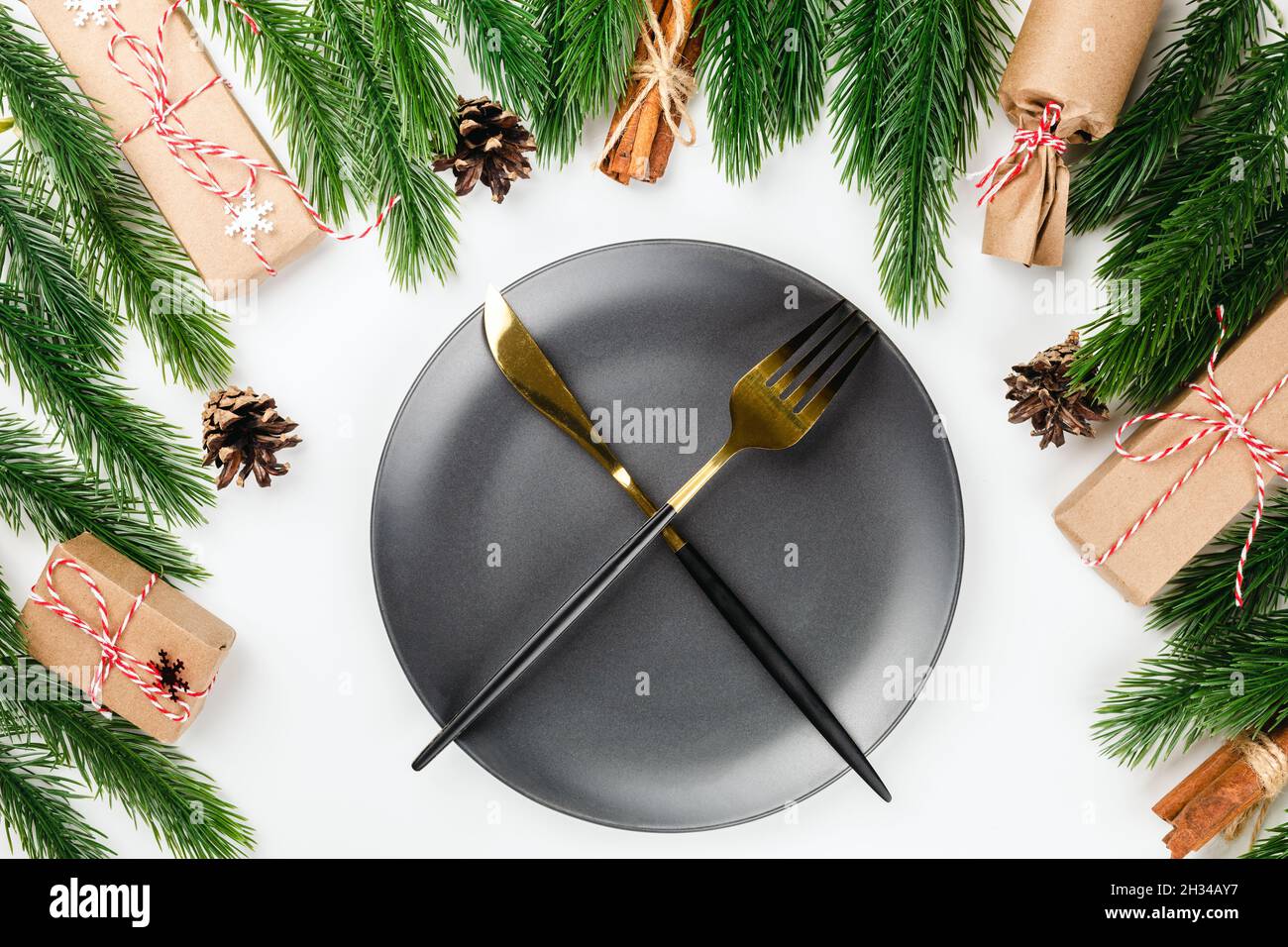 golden knife and fork crossed on black plate on Christmas dinner table with frame of fir branches, with cones and gifts Stock Photo