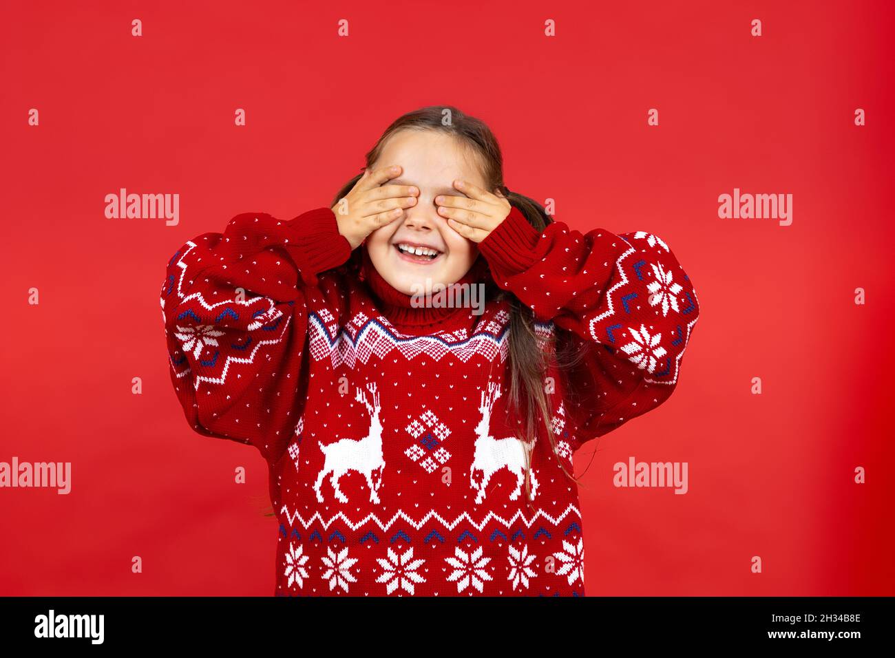 portrait of happy, cheerful charming girl in red Christmas sweater with reindeer covering eyes with palms and waiting for New Year miracle, isolated Stock Photo
