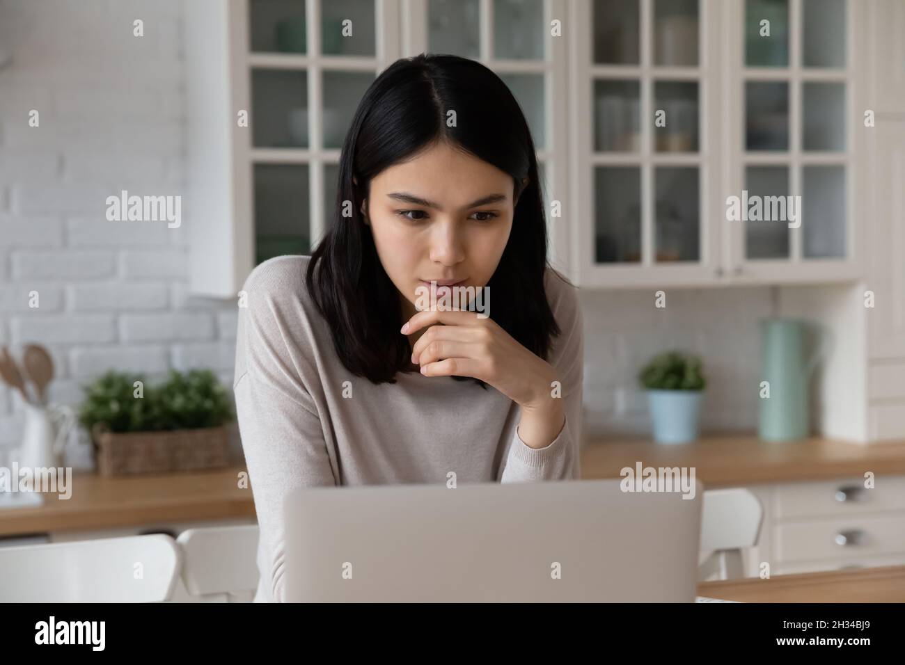 Serious focused Asian student using laptop in kitchen Stock Photo