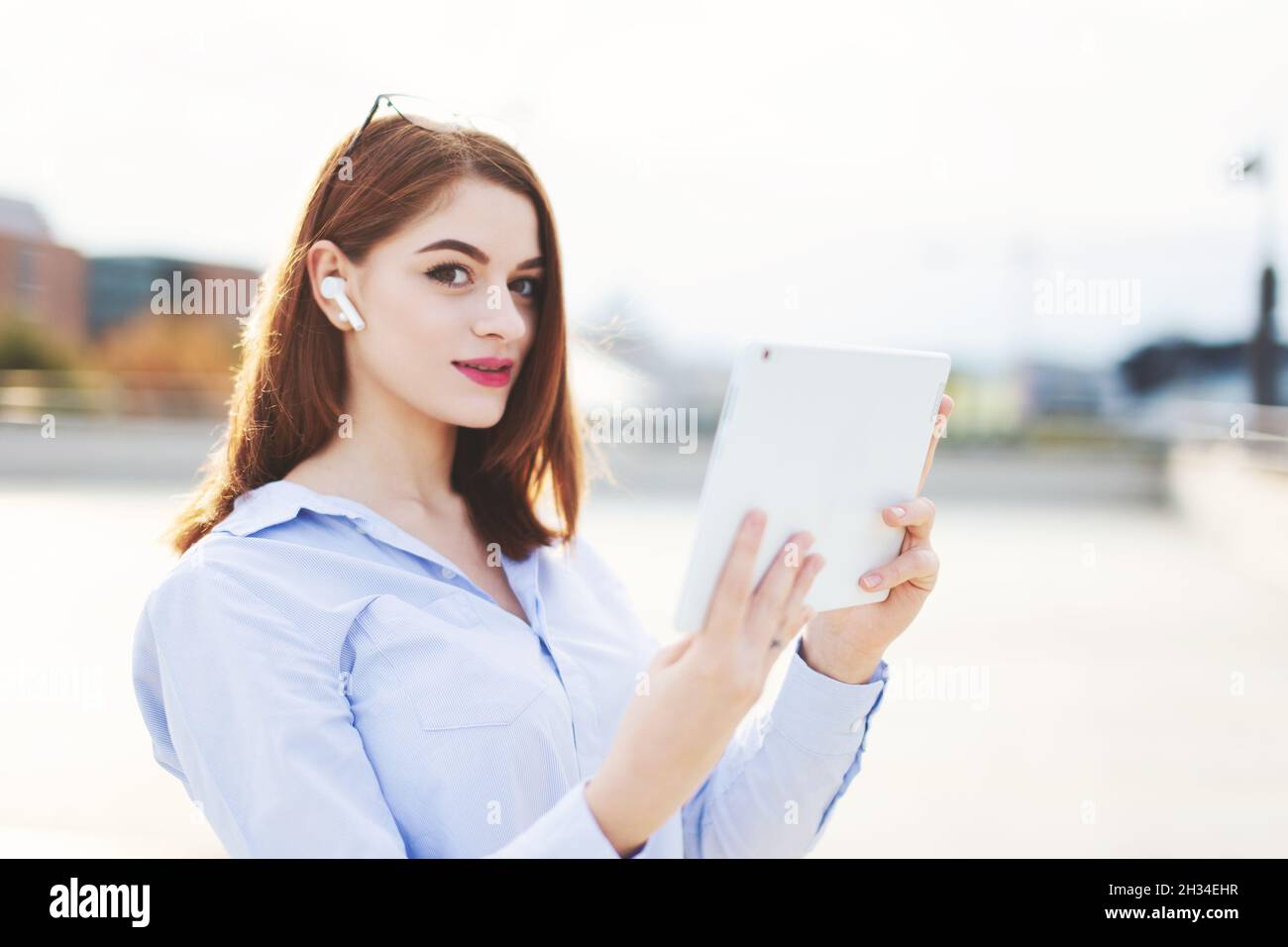 Happy young Caucasian redhead businesswoman with wireless headset using tablet outdoors Stock Photo