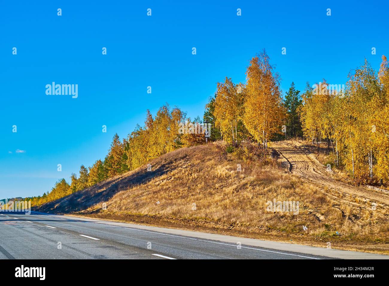 The small hill at the edge of intercity road with the autumn forest and the winding dirt road, Ukraine Stock Photo