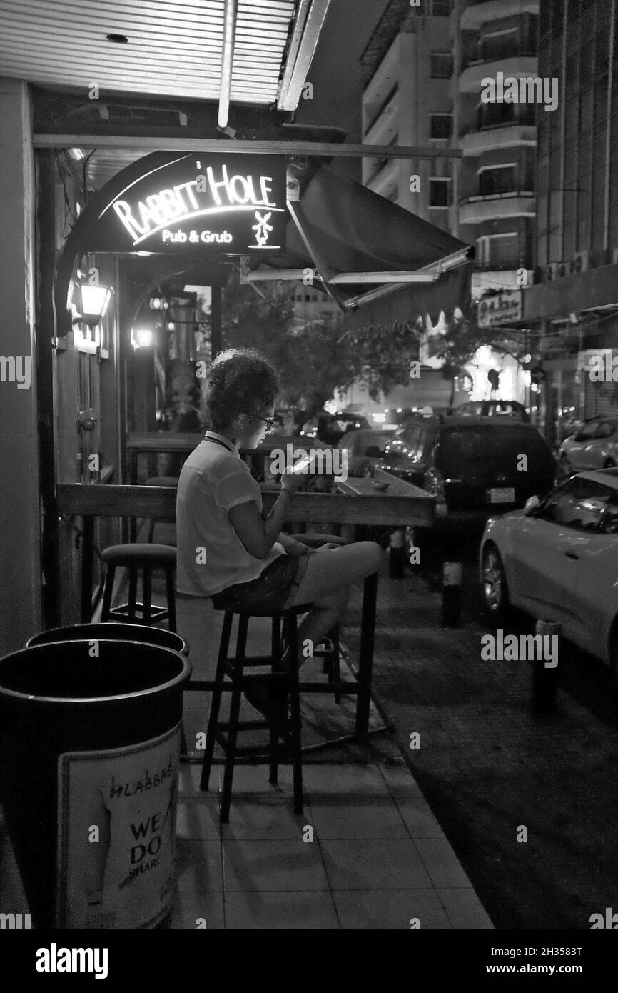 A young Lebanese woman uses her phone as sits on a stool outside The Rabbit Hole in the Hamra neighborhood in Beirut, Lebanon.  The area is filled with bars and restaurants catering to the large university population in the area. Stock Photo