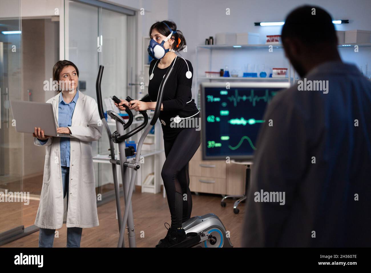 Specialist fitness researcher examining endurance controling EGK of athlete woman with mask running on bycle trainer. Physician man analyzing heart rate in medical modern laboratory. Medicine service Stock Photo