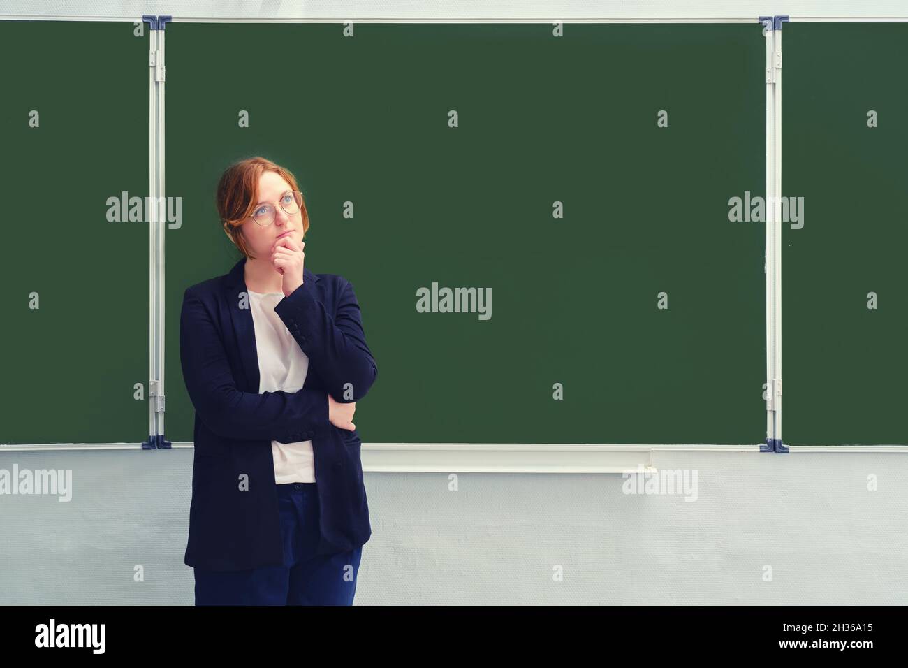A school teacher stands doubtfully in front of an empty blackboard, copy space for the inscription. Problems with learning via the Internet during the Stock Photo