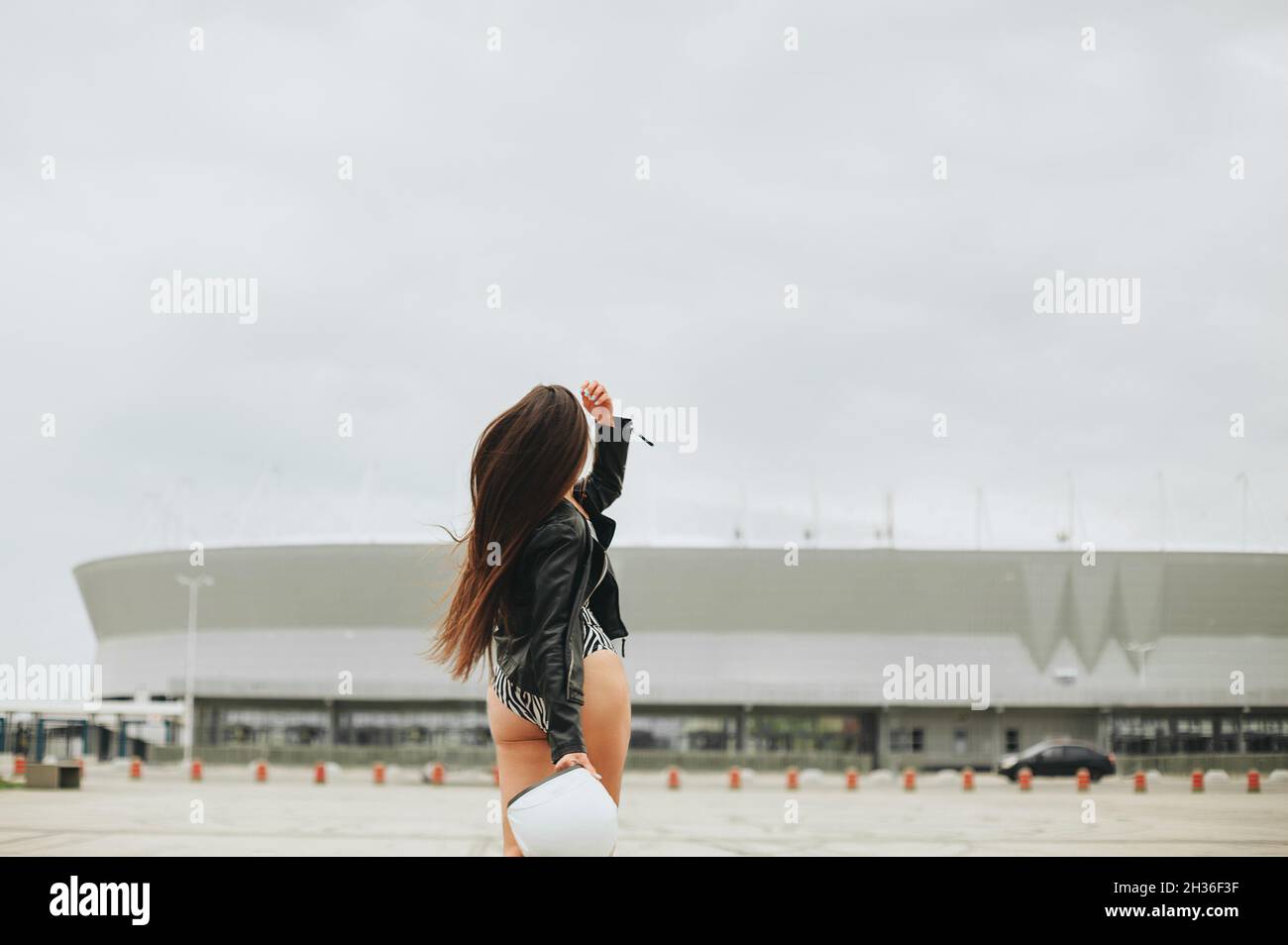 Rear view of slim brunette biker girl in bodysuit and leather jacket with safety bike helmet in hands posing at empty stadium outdoors Stock Photo