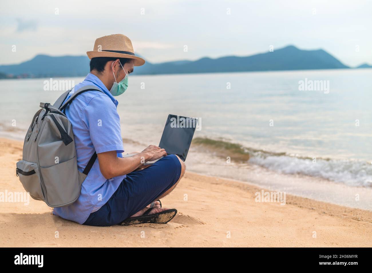 Man wear mask using laptop computer on beach  sea  and Man travel holiday Phuket sandbox Thailand are freedom life financial Stock Photo