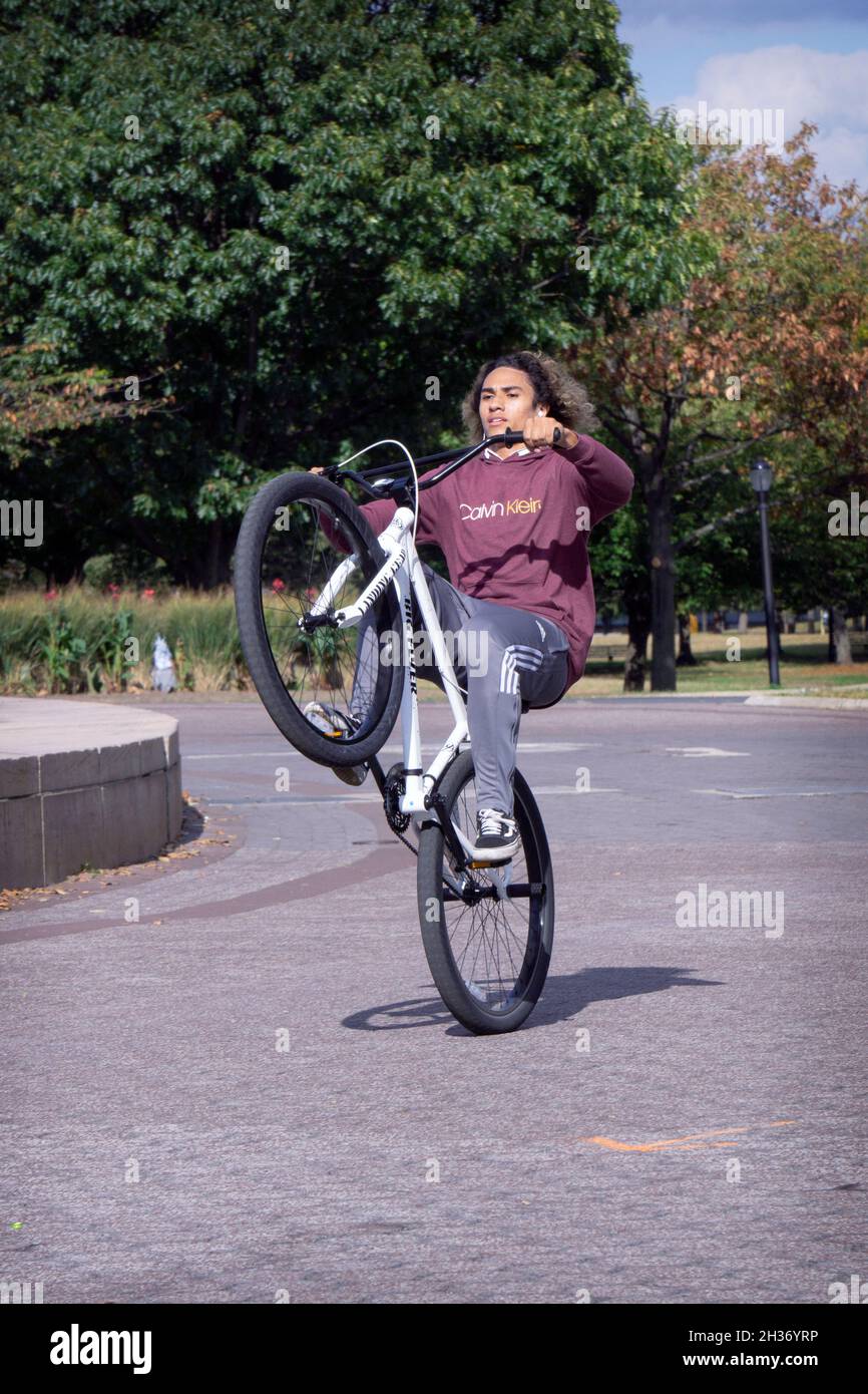 A young man with long hair does wheelies on his bike while riding laps around the Unisphere in Flushing Meadows Corona Park in Queens, New York. Stock Photo
