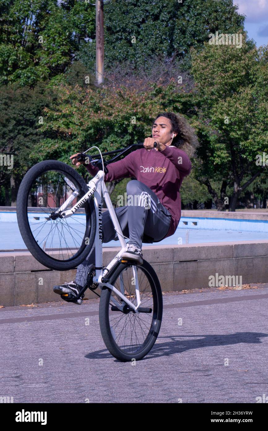 A young man with long hair does wheelies on his bike while riding laps around the Unisphere in Flushing Meadows Corona Park in Queens, New York. Stock Photo