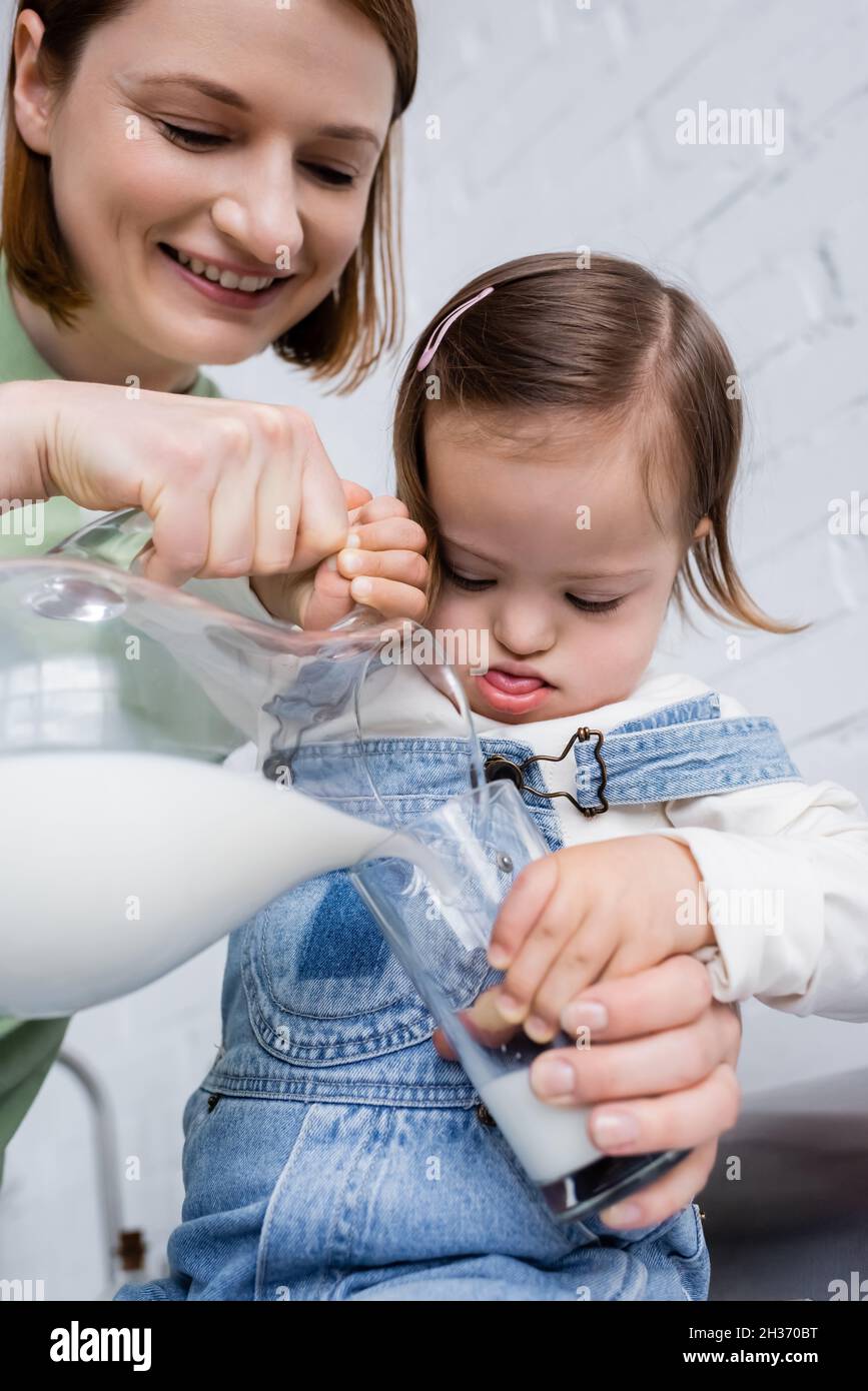 Smiling parent pouring milk from jug near kid with down syndrome in kitchen Stock Photo