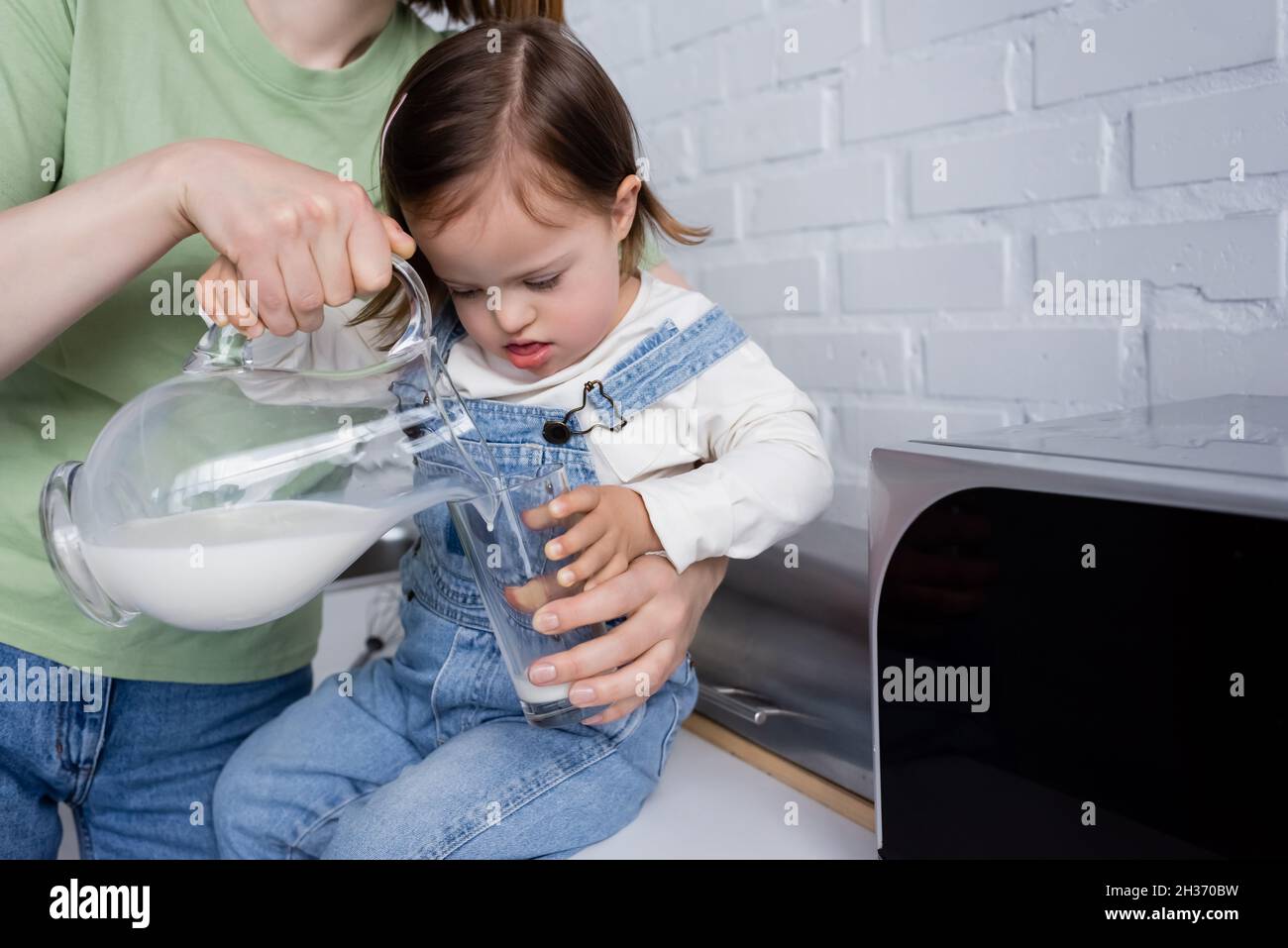 Woman pouring milk in glass near daughter with down syndrome in kitchen Stock Photo