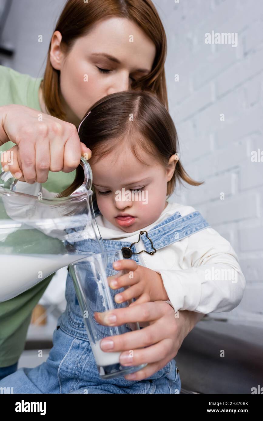 Woman pouring milk near kid with down syndrome in kitchen Stock Photo
