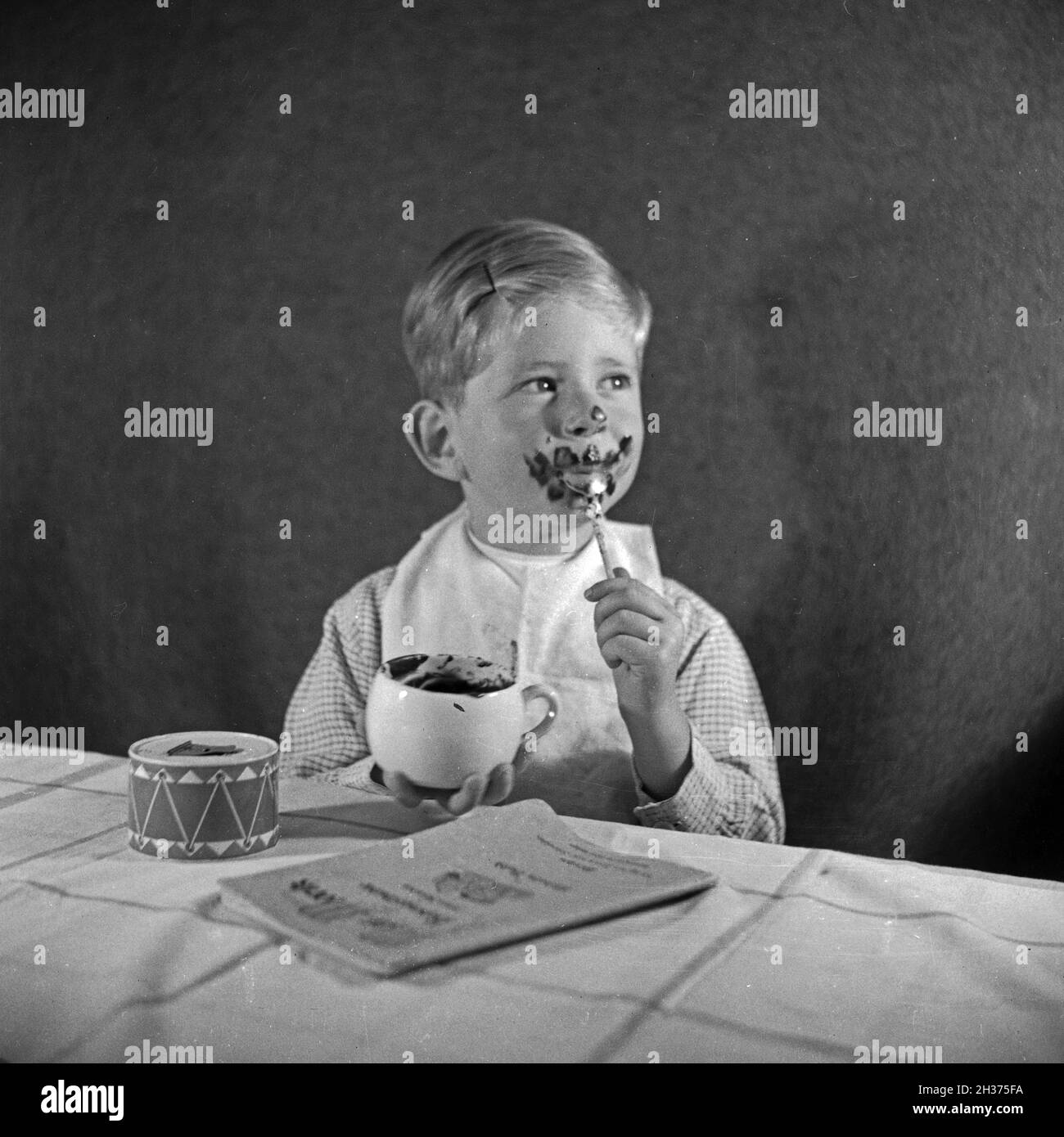 Ein kleiner Junge isst eine Tasse Schokoladenpudding und schmiert sich  damit richtig ein, Deutschland 1940er Jahre. A little boy eating a  chocolate pudding and is totally smeared, Germany 1940s Stock Photo -