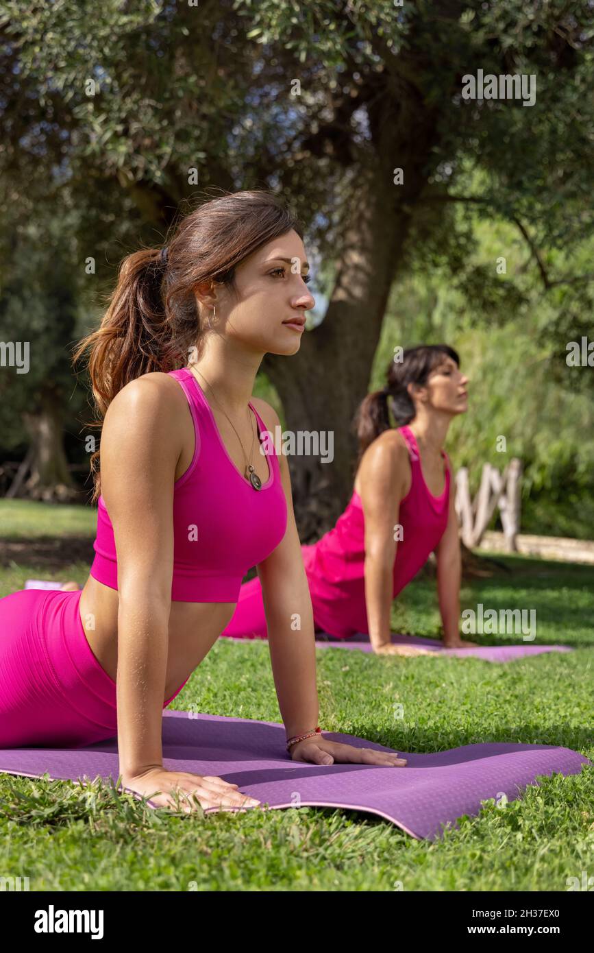 Two relaxed women practicing pilates pose Cobra in a park in a sunny day on summer. Young and middle-aged caucasian women in a pink clothing doing yog Stock Photo