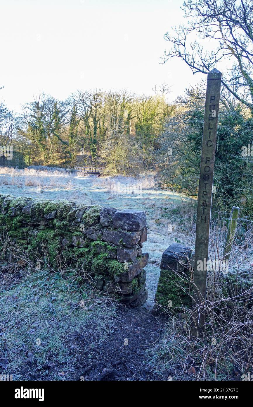 A wooden footpath marker and stone stile with the path leading to a wooden Bridge through a field covered in frost, Winter, Staffordshire, England, UK Stock Photo