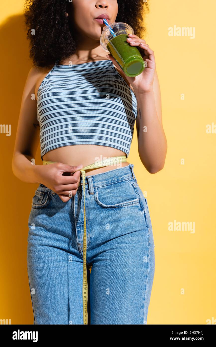 partial view of african american woman in striped tank top measuring waist while drinking smoothie on yellow Stock Photo