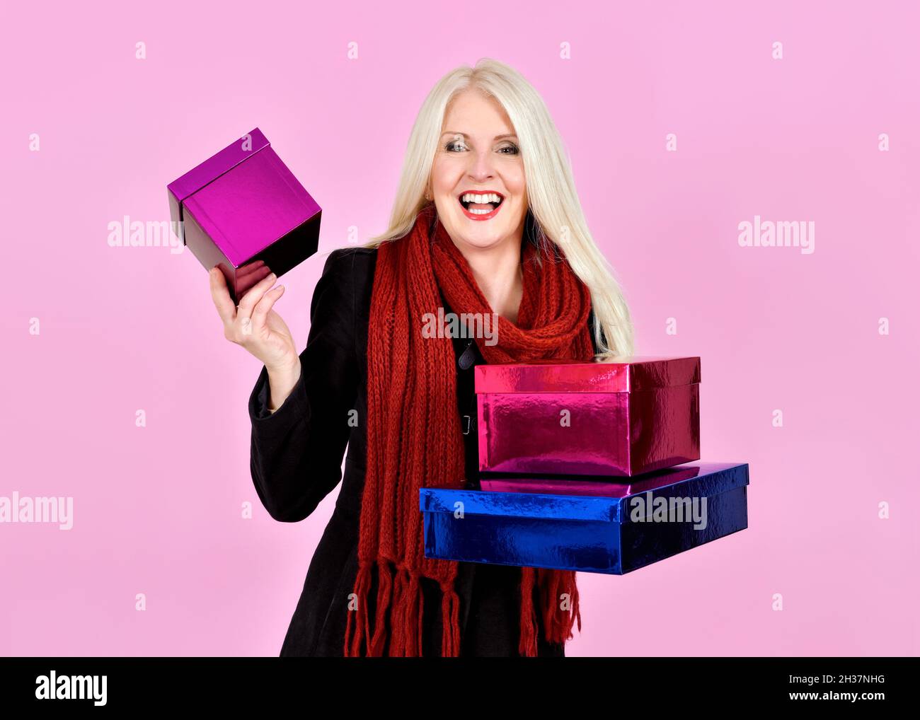 Attractive Blonde Lady dressed in coat and scarf laughing whilst carrying lots of coloured boxes or presents, taken on pink background Stock Photo