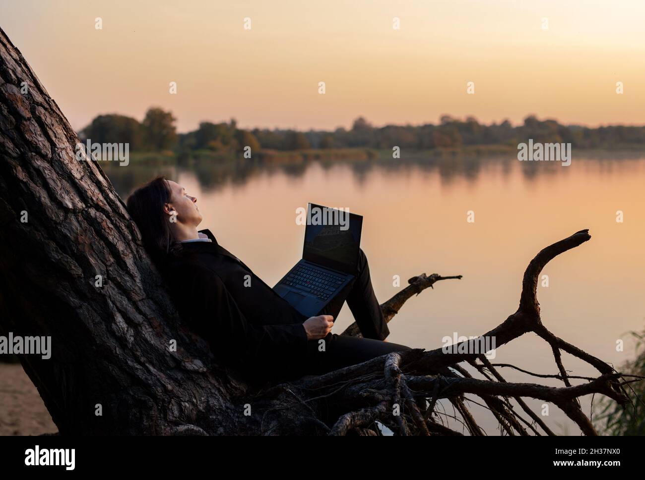 Handsome businessman in stylish black suit sits on tree trunk by the water with laptop in his hand at sunset.  Stock Photo