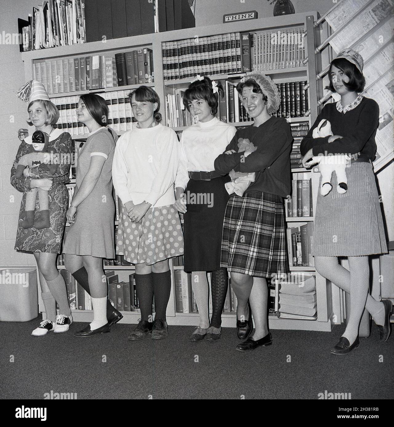 1960s historical, young female high school students in a study room or section of a library standing in a line, with three of them holding soft toys, with one girl on the left wearing a party hat and holding a batman doll, Virginia, USA. Possibly taking part in a some sort of class on imaginative play. Behind them, on top of a bookcase filled with textbooks, a sign saying THINK. Stock Photo