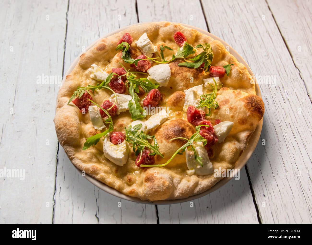 Pizza with mozzarella cheese, raw sausage and rocket in plate on white