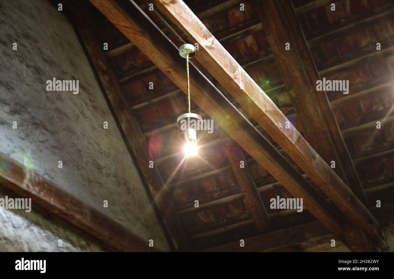 A glowing light bulb. An old lamp suspended from a wooden beam under the ceiling. Forgotten attic. Stock Photo