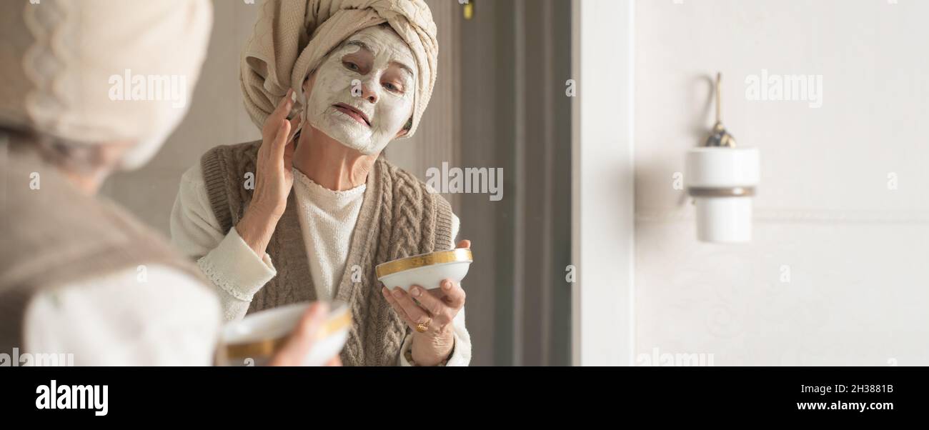 lderly woman with towel on her head smiles and applies cleansing mask . Stock Photo