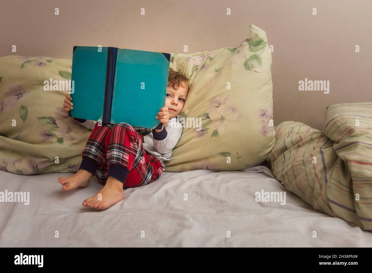 Toddler in pajamas sitting on bed and reading bedtime book looki Stock Photo