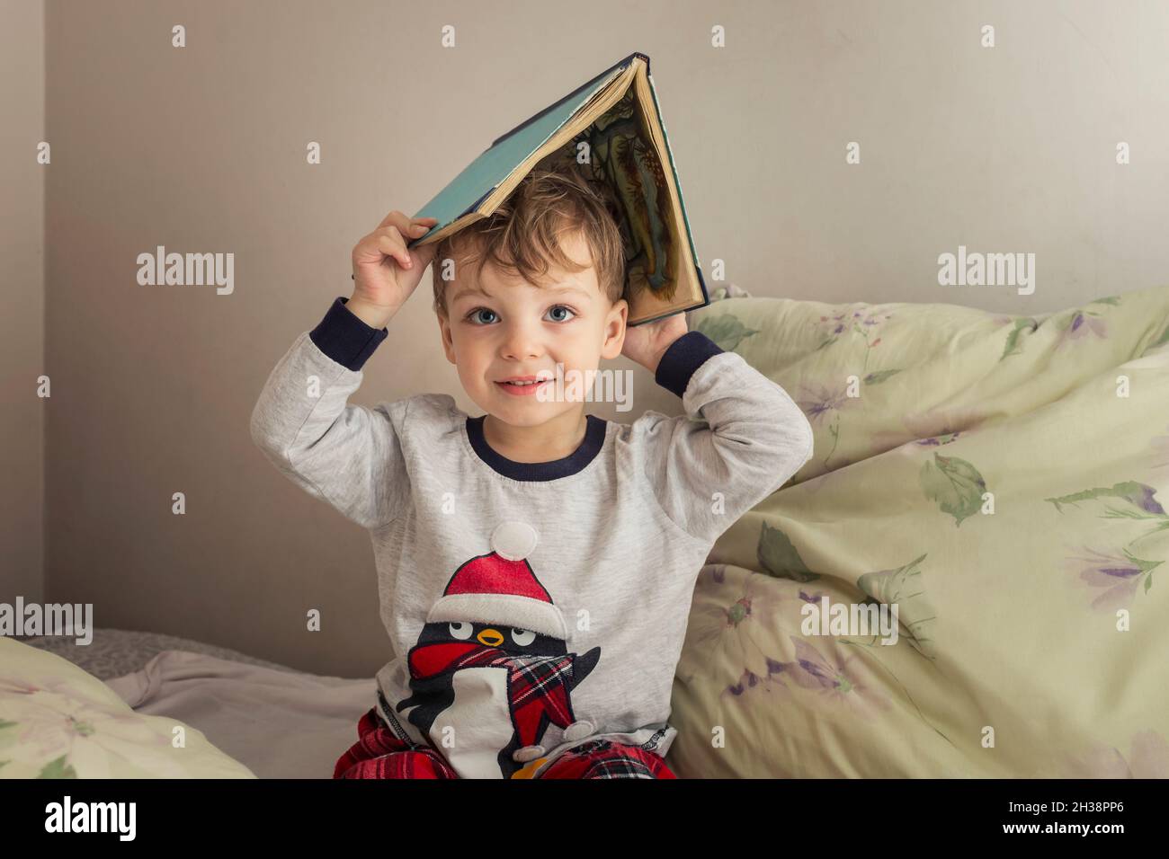 Toddler in pajamas putting book on his head and sitting on a bed Stock Photo