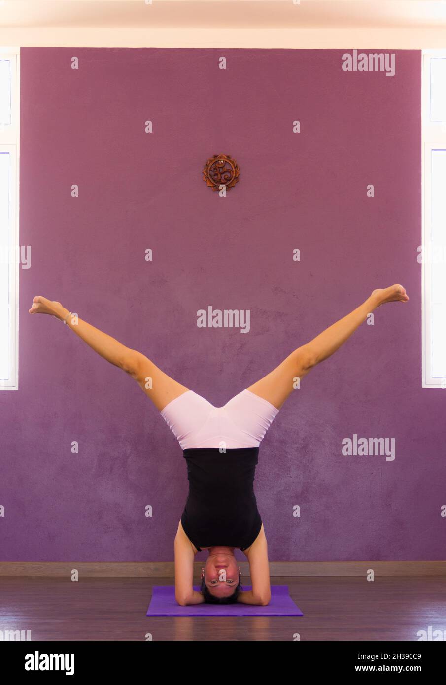 Young woman on headstand pose with split legs. Female yogi in sirsasana on purple wall studio. Balance, equilibrium, flexibility, exercise, workout Stock Photo