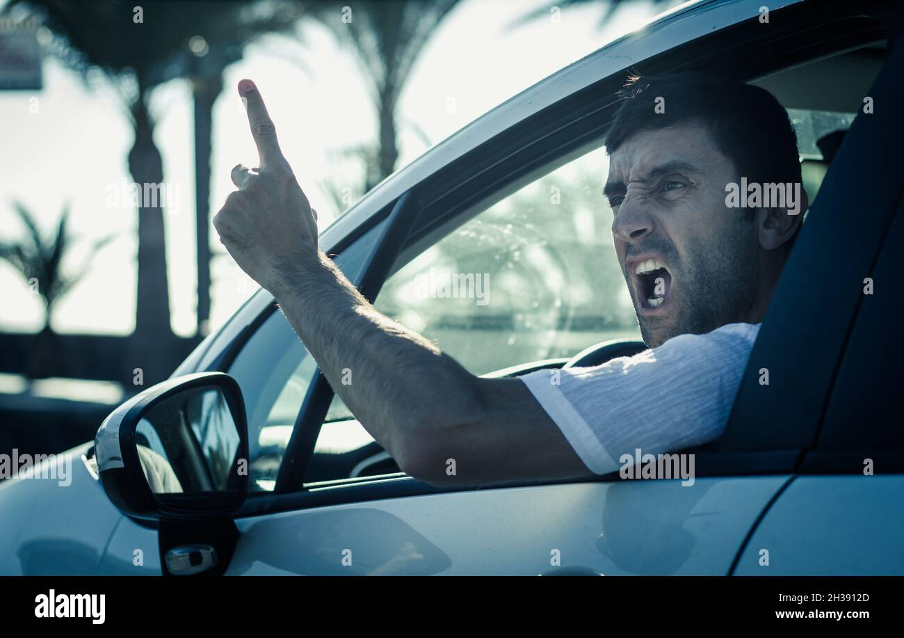 Furious man gesticulates with hand out of window while driving. Crazy driver yelling from car at sunrise. Stress, traffic rush hour, anxious, madness Stock Photo