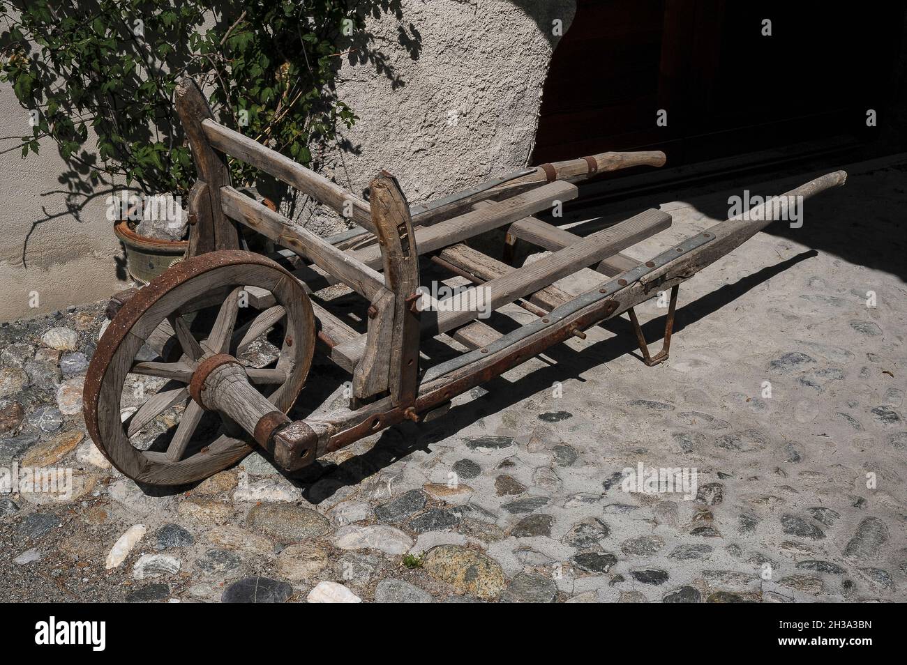An antique or vintage wooden sack truck or sack barrow, with a timber axle and a single wheel bound by an iron tyre, rests on a sunlit cobbled path in the historic Romansh-speaking village of Ardez, in the Lower Engadine Valley in Graubünden or Grisons canton, eastern Switzerland. Stock Photo