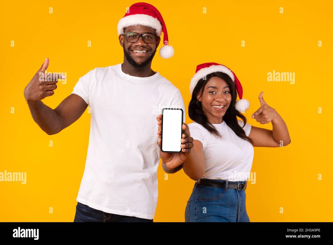 Black Couple Wearing Santa Hats Showing Cellphone Screen, Yellow Background Stock Photo