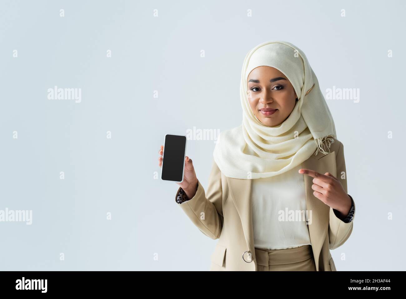 happy muslim woman in hijab pointing at smartphone with blank screen ...