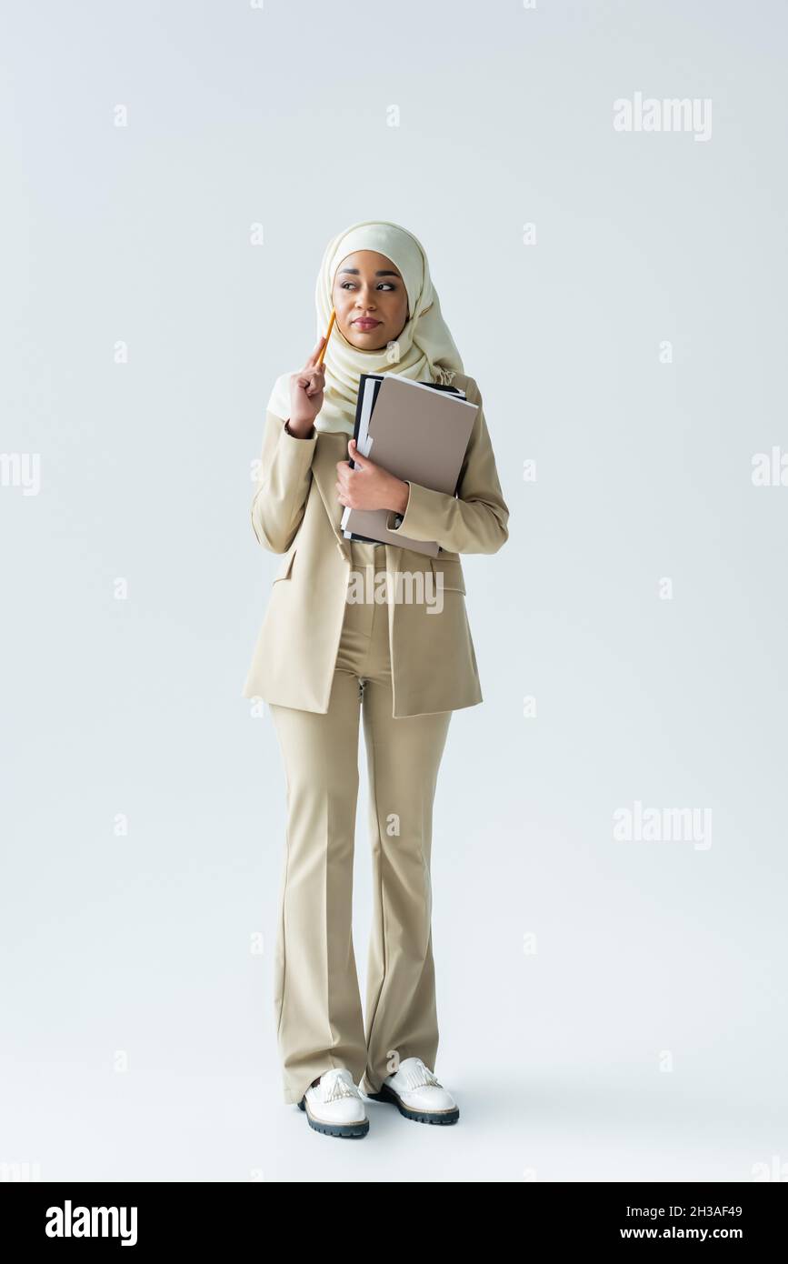 full length of pensive muslim woman in hijab holding pencil and folders ...