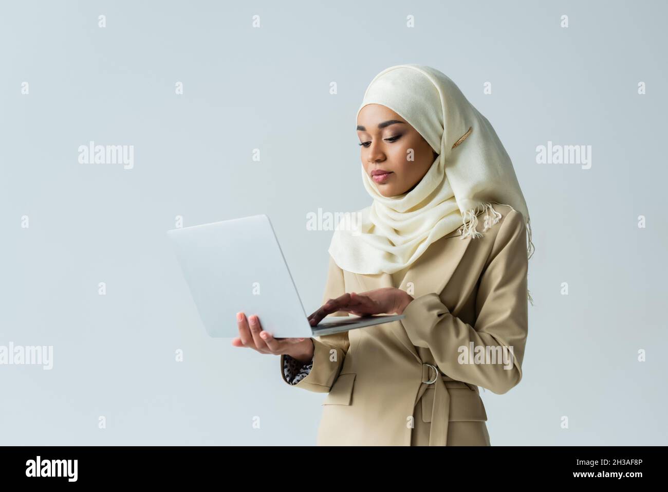 stylish muslim woman in hijab and beige suit typing on laptop isolated ...