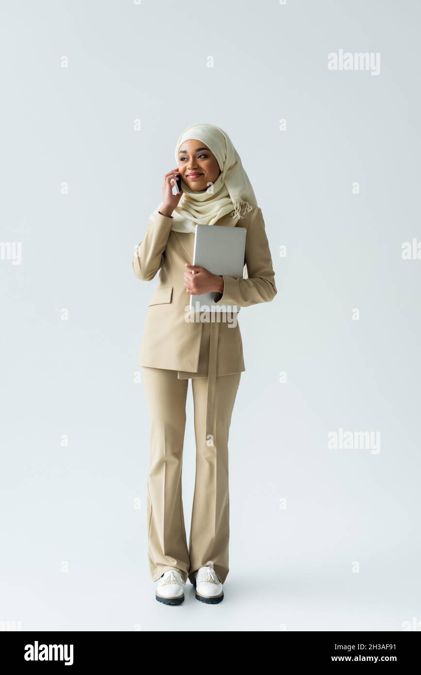 full length of happy muslim woman in hijab and beige suit talking ...