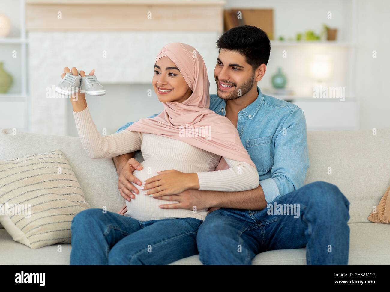 Loving arab man hugging his pregnant wife with baby booties, sitting together on sofa at home Stock Photo