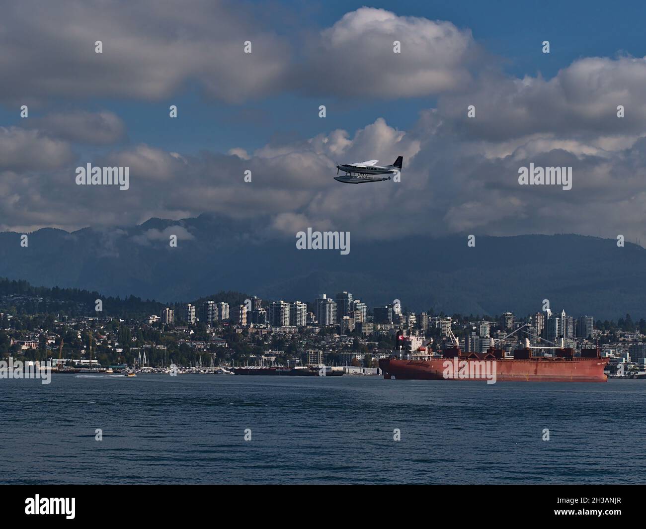 Red bulk cargo vessel lying at anchor in Burrard Inlet in front of North Vancouver downtown with seaplane passing by and mountains in background. Stock Photo