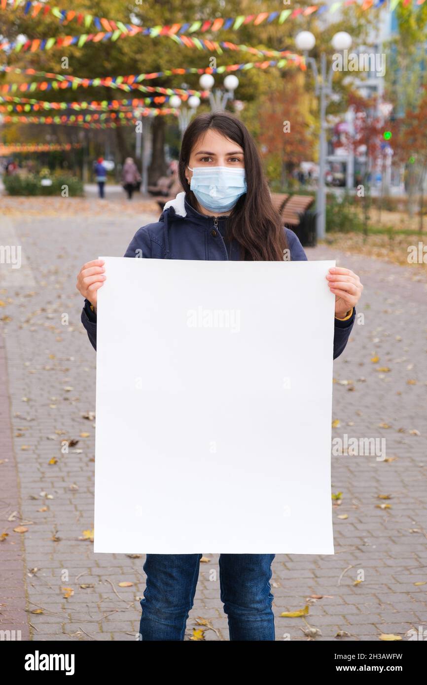 A woman in a protective mask holds a vertical sheet of paper without text Stock Photo