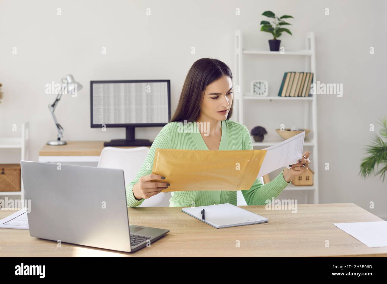 Young woman holding envelope and reading letter while sitting at desk with laptop at home Stock Photo